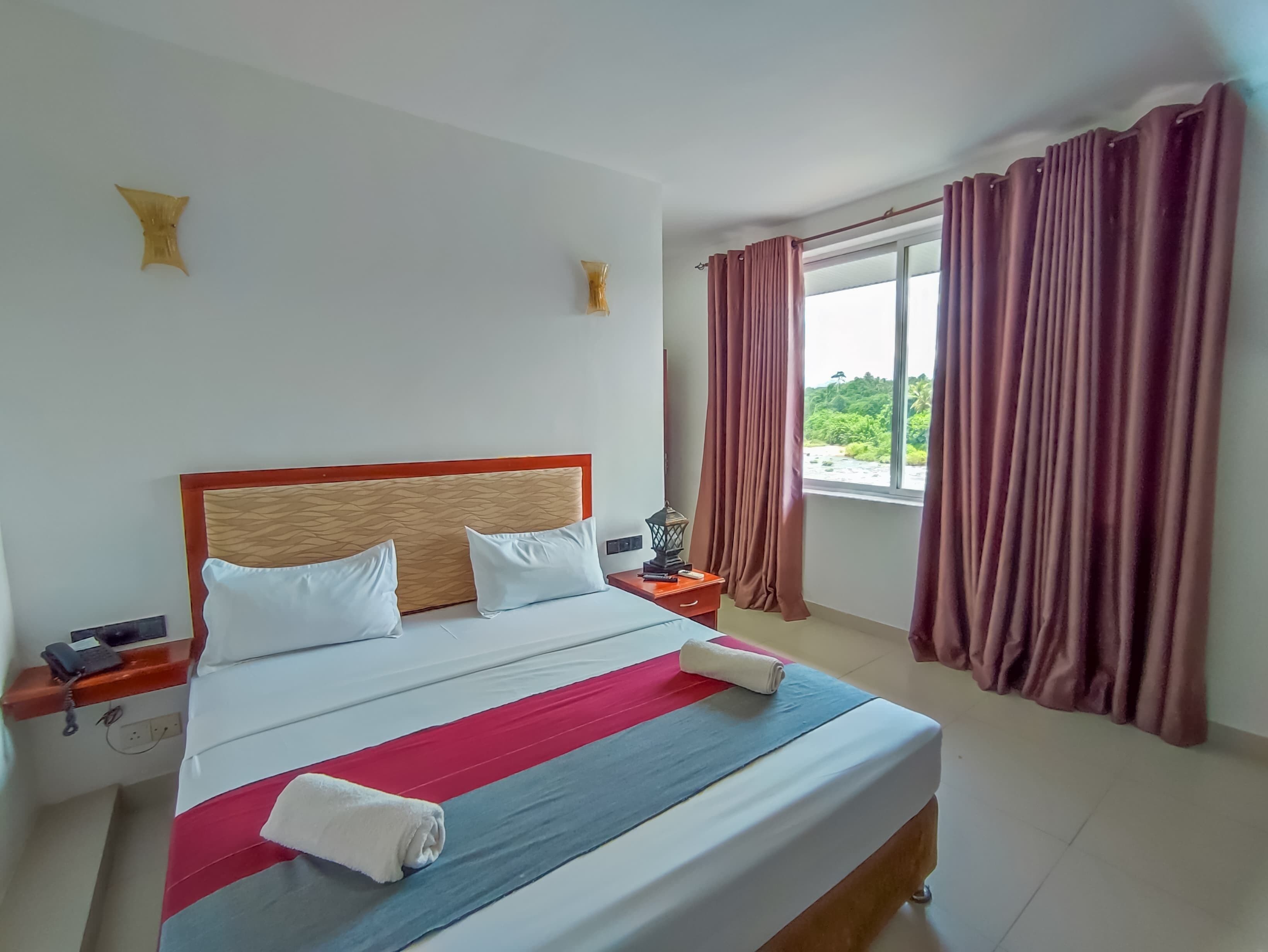 Ein ordentlich bezogenes Bett mit einer gestreiften Tagesdecke, zwei Kissen und einem zusammengerollten Handtuch. Ein Nachttisch mit Lampe und Telefon ist vorhanden. Das Zimmer hat ein großes Fenster mit Vorhängen und Blick auf Grünfläche.