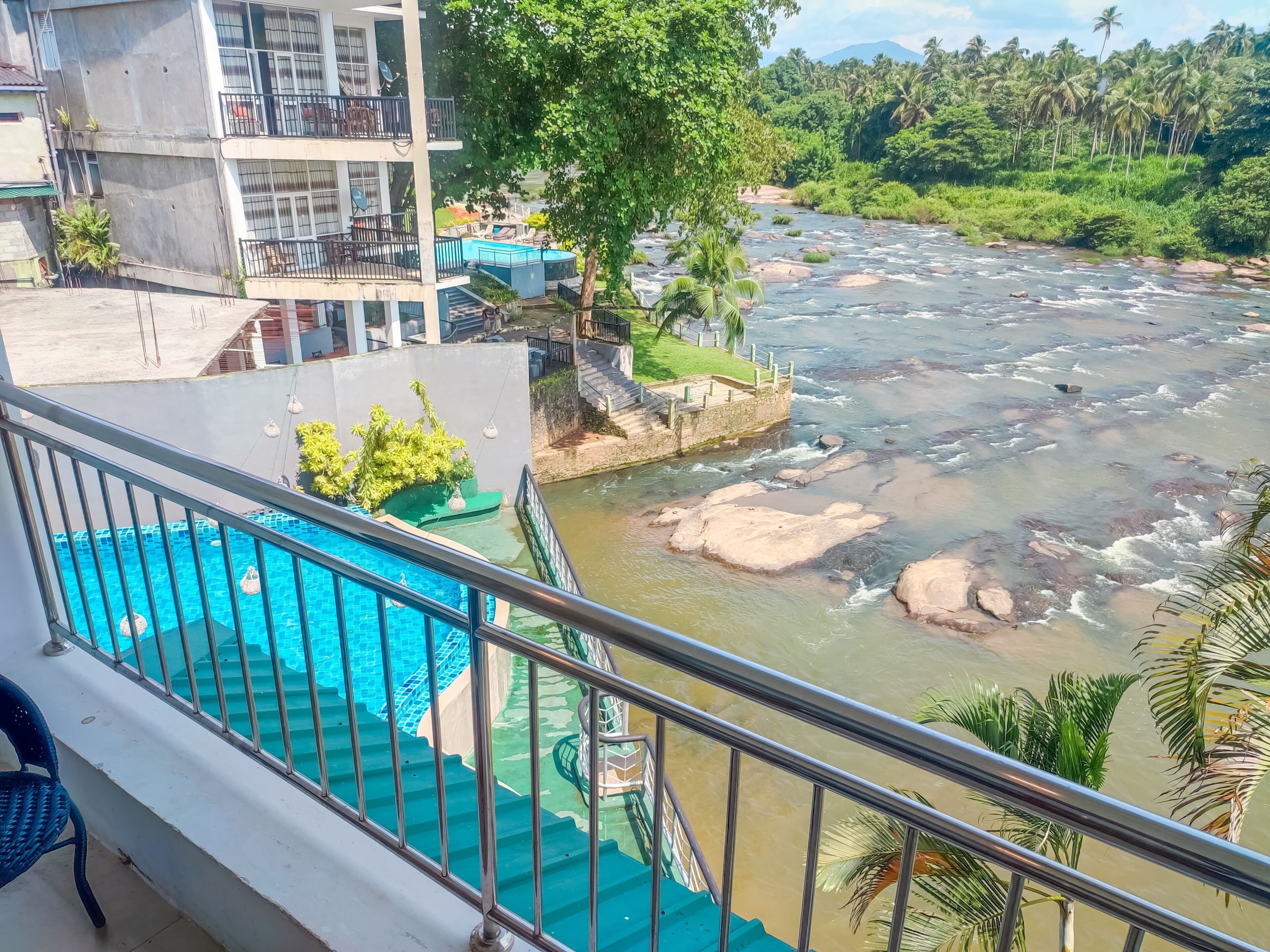 Ein Hotel mit Pool, einer Treppe, die zu einem Balkon mit Flussblick führt, und Blick auf Palmen und Grünpflanzen.
