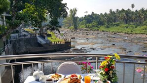 Een ontbijttafel met brood, fruit en thee, met uitzicht op een rivier met een brug en weelderige begroeiing.