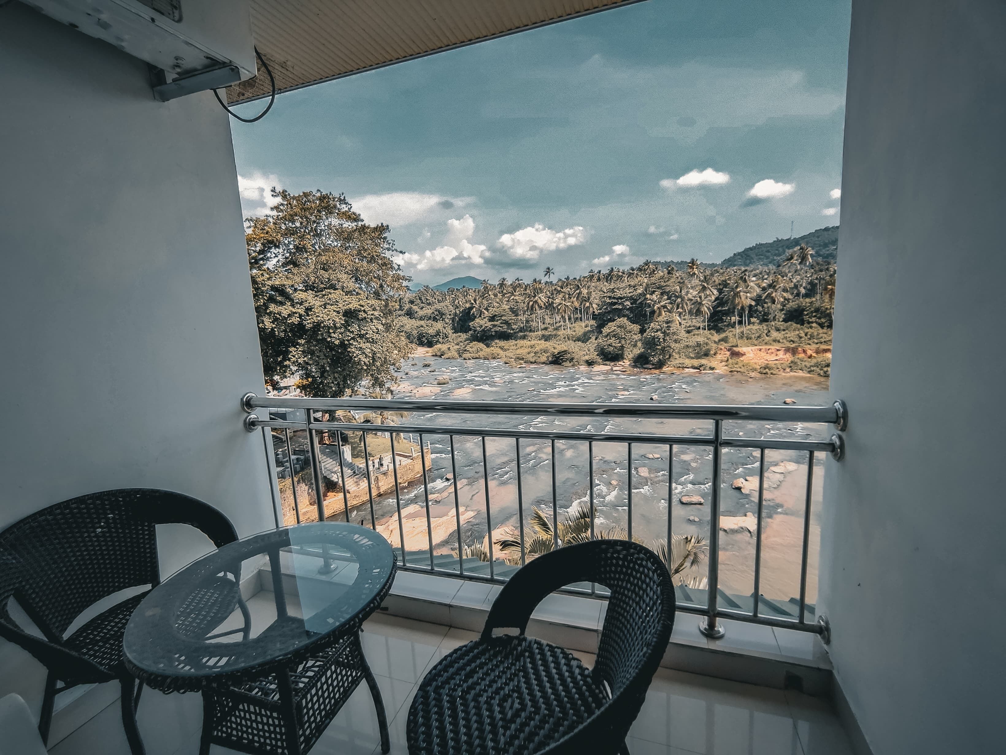 A balcony with two wicker chairs and a glass table, overlooking a river with a mountainous landscape and palm trees in the background.
