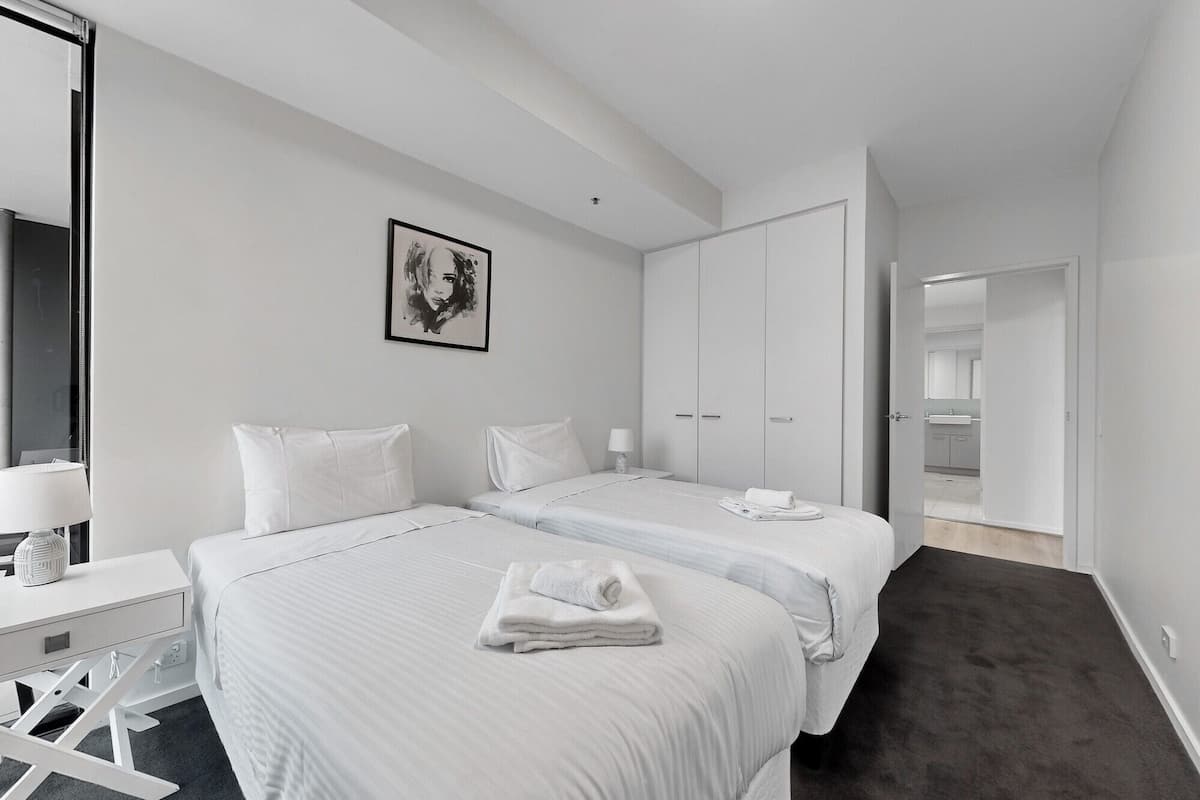 A hotel room with two beds, a desk, a chair, a wardrobe, and a bathroom visible through an open door.