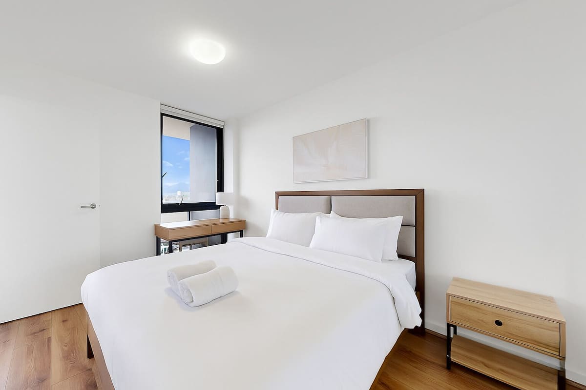 A modern hotel room with a large bed, a bedside table, a wooden nightstand, and a window with a view.