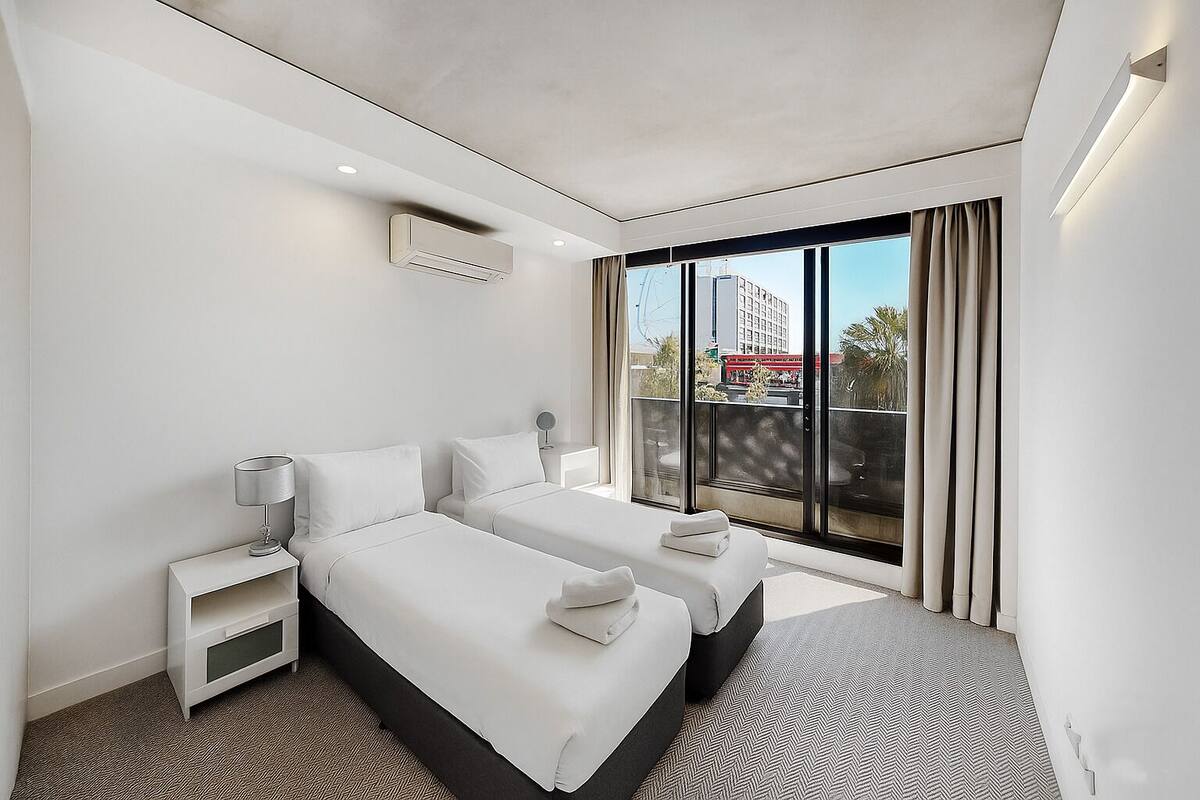 A modern hotel room with two beds, a large window with a view of the city, and a small bedside table with a lamp.