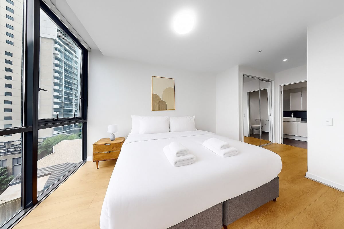 A modern hotel room with a large bed, a bedside table with a lamp, a view of the city through large windows, and a door leading to a bathroom.