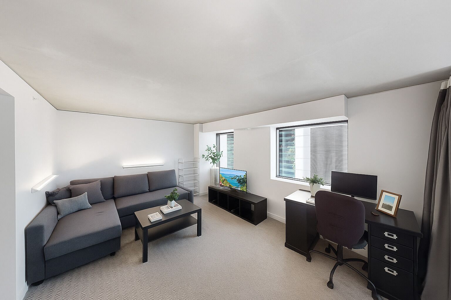 Un salon moderne avec un canapé gris, une table basse, un téléviseur, un bureau avec une chaise et une fenêtre avec des stores.