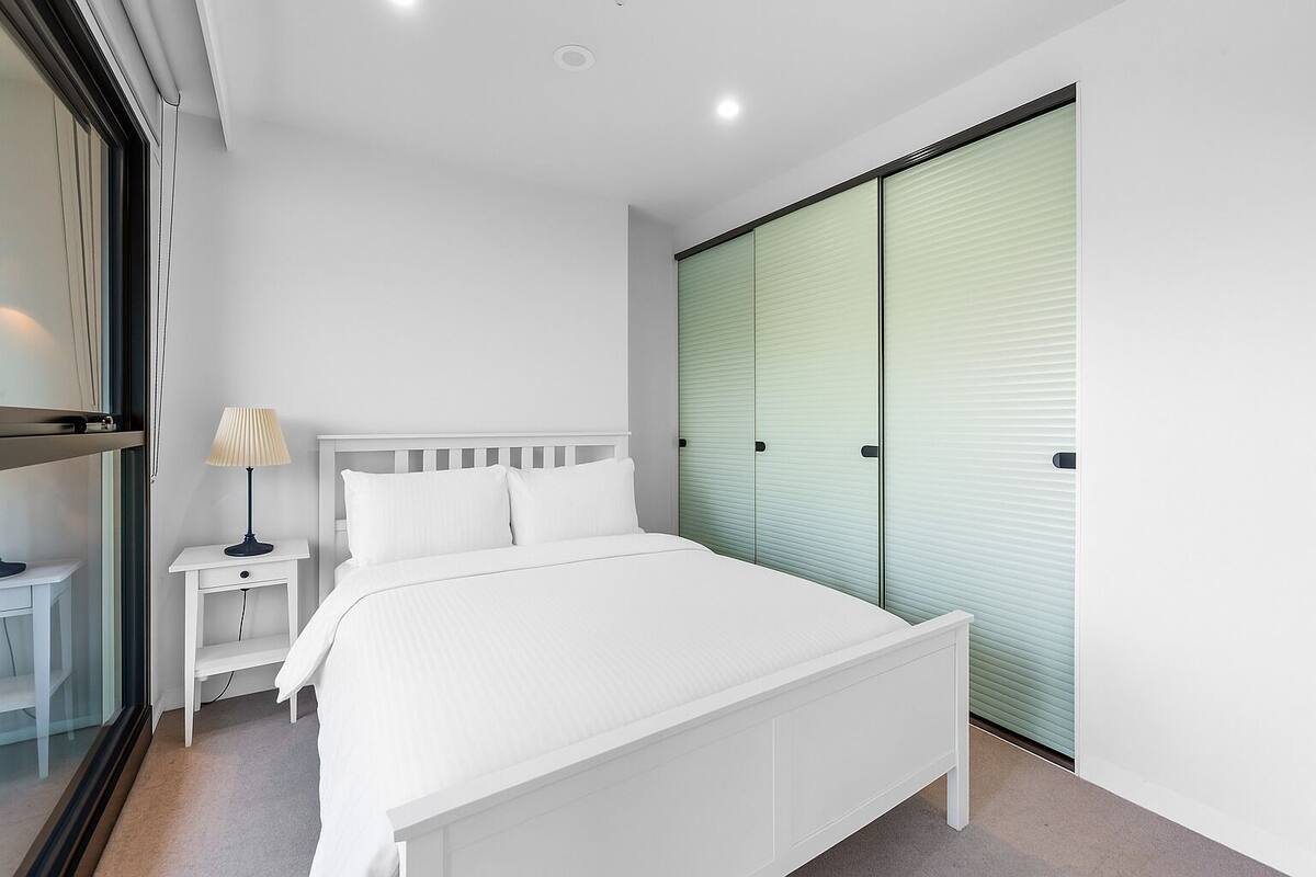 A modern bedroom with a white bed, a nightstand with a lamp, and a sliding glass door closet.