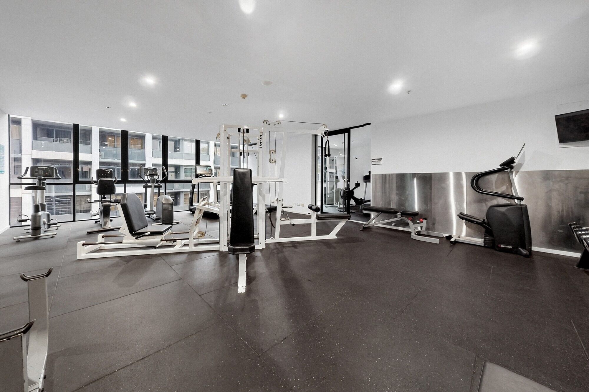 Een moderne sportschool met diverse fitnessapparaten, een grote spiegel en uitzicht op een gebouw door middel van grote ramen.