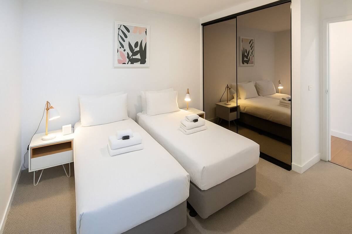 A modern hotel room with two single beds, a bedside table with a lamp, and a sliding door leading to a larger room.