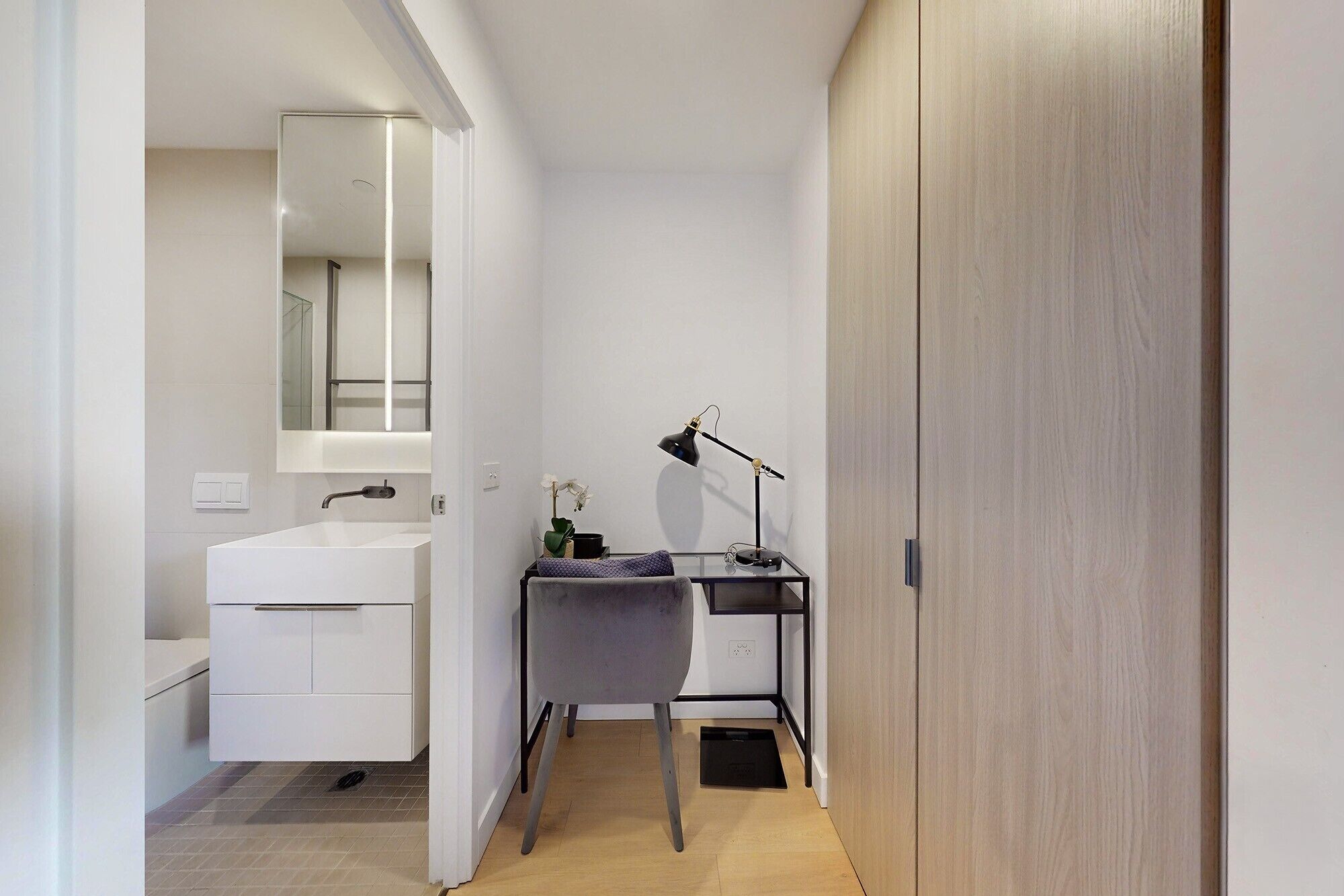 A modern bathroom with a white sink, mirror, and bathtub. There is a desk with a lamp and a small plant. The room has wooden flooring and white walls.