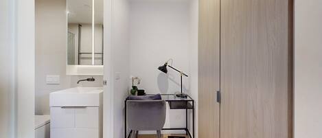 A modern bathroom with a white sink, mirror, and bathtub. There is a desk with a lamp and a small plant. The room has wooden flooring and white walls.