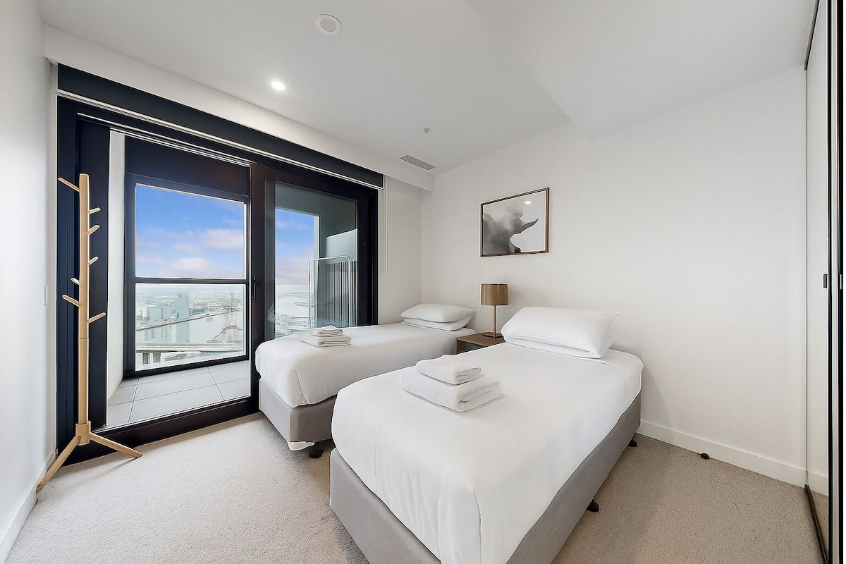 A modern hotel room with two single beds, a large window with a city view, a bedside table with a lamp, and a framed artwork on the wall.