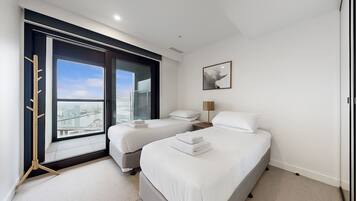 A modern hotel room with two single beds, a large window with a city view, a bedside table with a lamp, and a framed artwork on the wall.