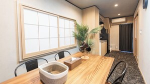 A dining area with a wooden table, chairs, a large window with a grid pattern, a potted plant, and a kitchen area in the background. - b&Kuromon (Osaka)