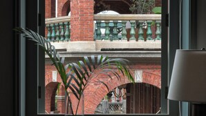 Vista attraverso una finestra su un edificio in mattoni con archi, un balcone con balaustre verdi e un tavolo con un vaso e un piatto di cibo.