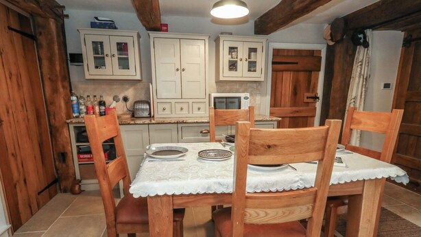 Une table de salle à manger en bois, accompagnée de quatre chaises, une nappe en dentelle blanche et un coin cuisine doté de meubles en bois et d’un four à micro-ondes.