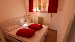 A single bed with white bedding and red pillows, a bedside table with a lamp, a radiator, and a window with floral curtains. - Quiet cozy apartment overlooking the lake, closed parking lot and pool, (Malcesine)