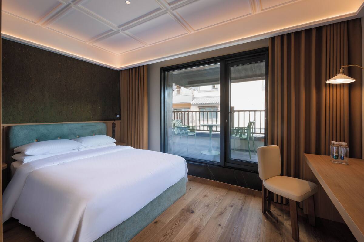 A hotel room with a large bed, a desk, a chair, and a view of the outside balcony.