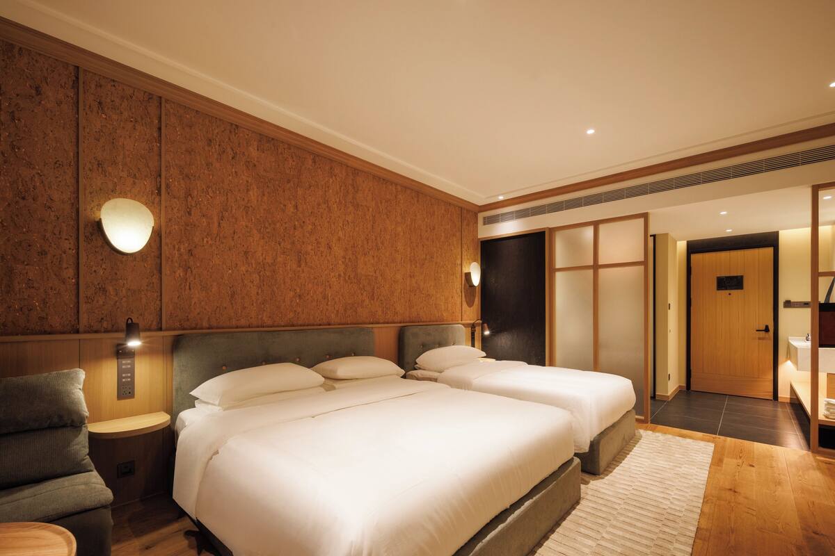 A hotel room with two beds, a nightstand, a wall-mounted light, and a wooden floor.