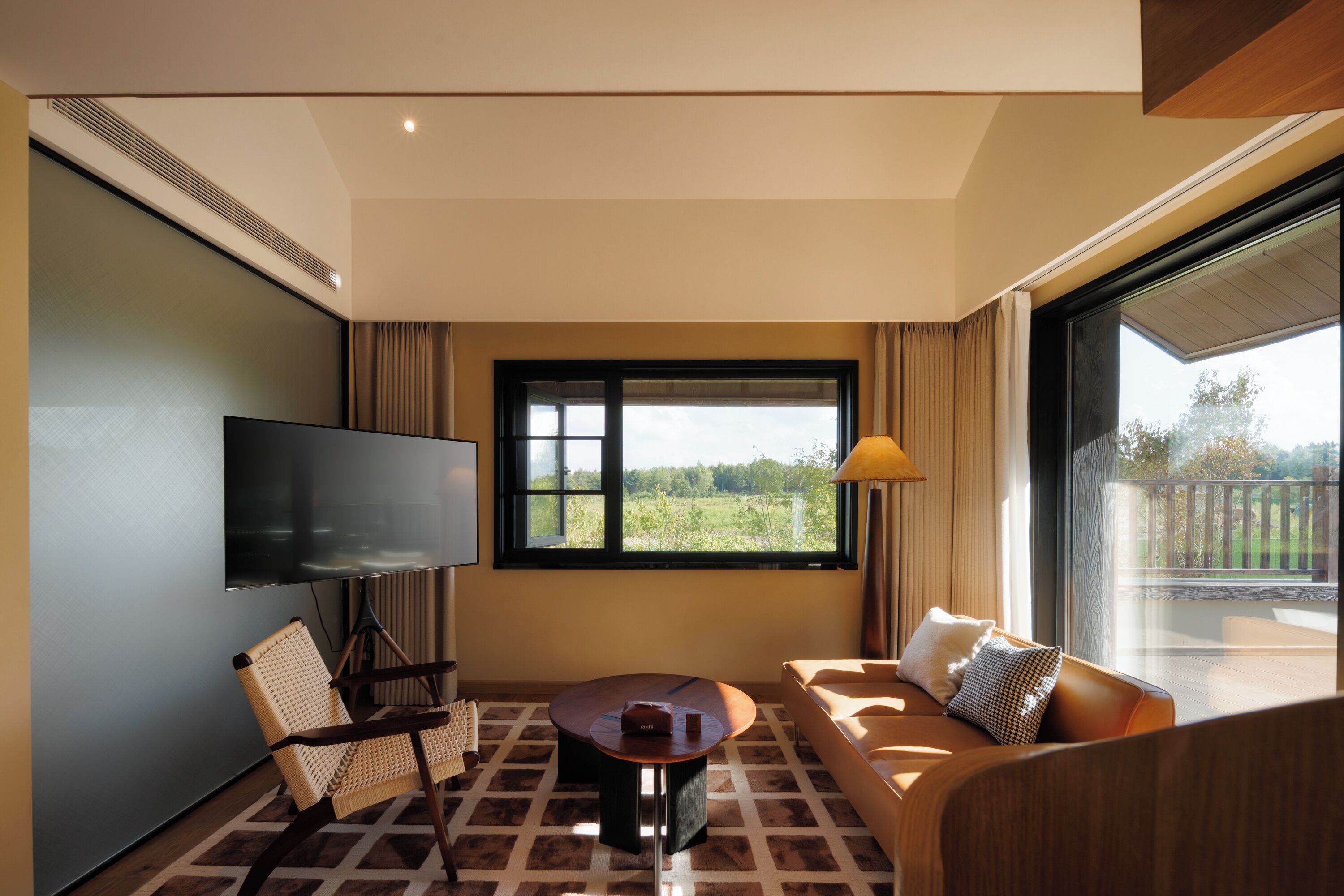 A modern living room with a flat-screen TV, a round coffee table, a sofa, and a large window offering a view of greenery.
