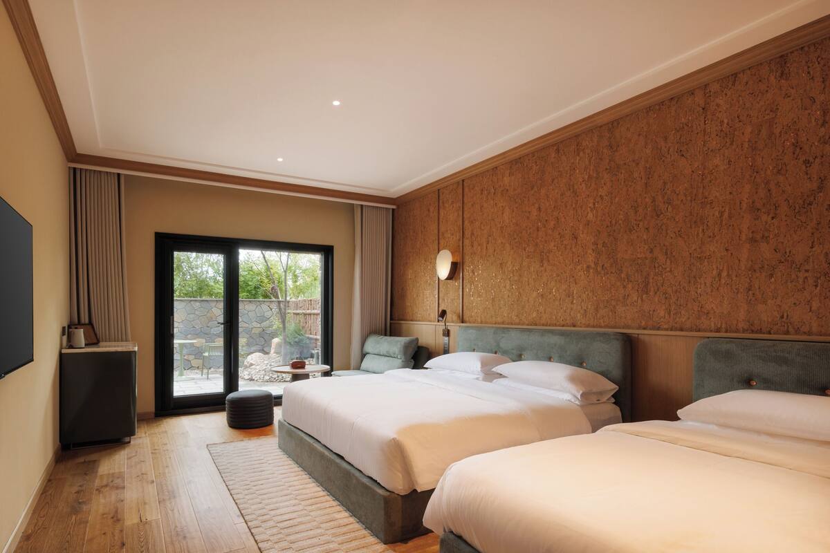 A hotel room with two beds, a flat-screen TV, a stone wall, and a view of the outdoors.