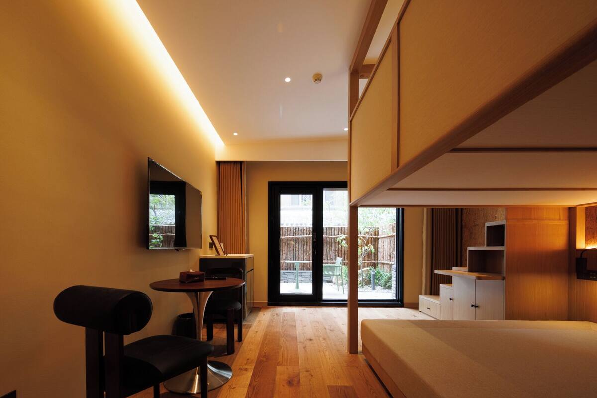 A modern hotel room with a bed, a desk, and a chair. There is a television mounted on the wall and a window with a view of the outside.