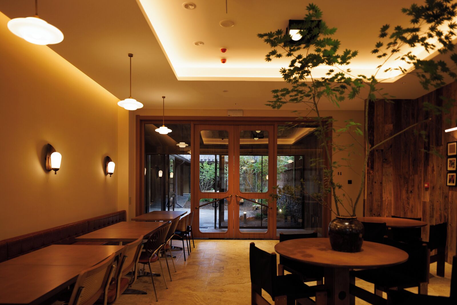 Un restaurant avec des tables et des chaises en bois, des luminaires suspendus et une grande porte-fenêtre coulissante donnant sur un espace extérieur.