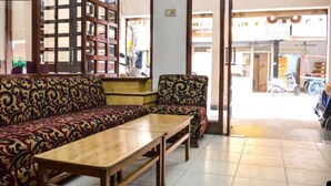 A seating area with patterned sofas and a wooden table, a wooden bookshelf, and a view of the street outside. - Hotel Saiom Palace (Raipur)