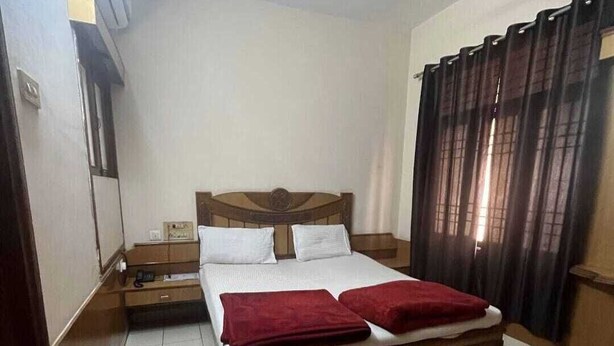 A bedroom with a wooden bed, white bedding, a nightstand, a red blanket, and a window with curtains. - Hotel Saiom Palace (Raipur)