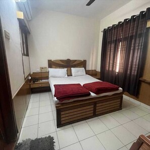 A bedroom with a wooden bed, white bedding, a nightstand, a red blanket, and a window with curtains. - Hotel Saiom Palace (Raipur)