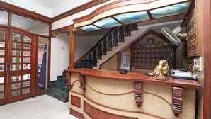 A reception desk with a wooden counter, a staircase with black railings, and a fan on the wall. - Hotel Saiom Palace (Raipur)