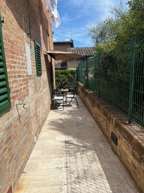 A narrow outdoor corridor with a brick wall, a green metal fence, and a table with chairs.