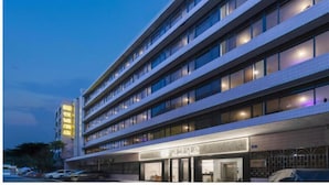 Ein mehrstöckiges Hotel mit modernem Design, das eine markante Eingangszone mit einem Schild, zahlreiche Fenster in jeder Etage und einen Parkplatz davor aufweist.