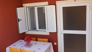 Uma área de refeições com uma mesa posta para dois, um vaso de flores e uma janela com persianas.
