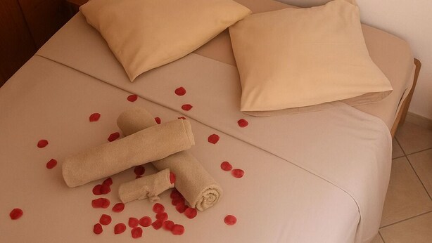 Cama bem arrumada com lençóis e almofadas bege, uma toalha enrolada e pétalas de rosa espalhadas.
