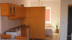 Cozinha compacta com armários de madeira, fogão e mesa de jantar com cadeiras. Há uma cama pequena e um vaso de planta no canto.