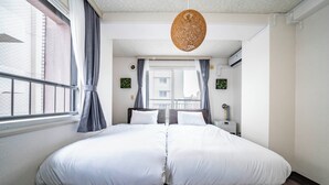 Ein Doppelbett mit weißer Bettwäsche, eine hängende Lampenschirm aus Rattan und Blick auf Gebäude durch ein Fenster mit grauen Vorhängen.