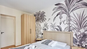Une chambre avec un grand lit, une armoire en bois, une petite table ronde et une fresque murale représentant des plantes tropicales.