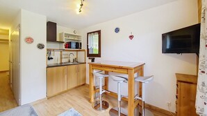 A compact kitchen with a microwave, sink, and wooden cabinets. There is a small dining area with a table and two bar stools. A flat-screen TV is mounted on the wall. - Toison Blanche TBC45 COSY & Mountain 4 Pers (Châtel)
