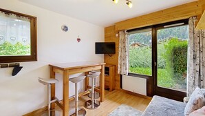 A room with a wooden table, bar stools, a TV, and a large window with a view of greenery. - Toison Blanche TBC45 COSY & Mountain 4 Pers (Châtel)