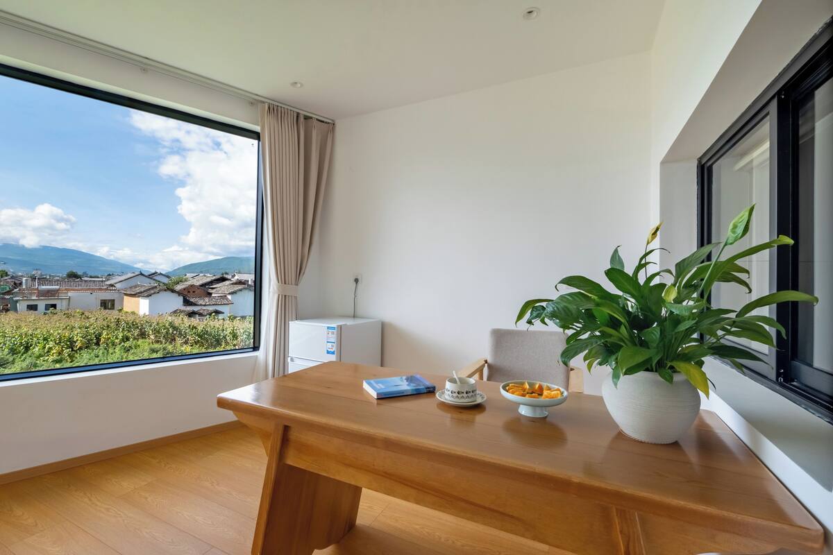 Een kamer met een houten bureau, een bloempot, een schaal fruit en uitzicht op huizen en bergen door een groot raam.