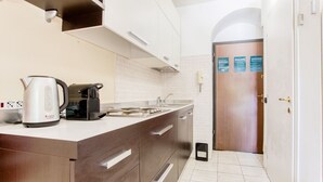 A modern kitchen with a built-in stove, a kettle, and a coffee maker. There is a sink with a faucet and a door with a sign in the background. - Easylife - Studio with balcony in Porta Romana (Milano)