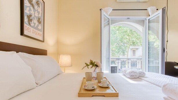 A hotel room with a bed, a tray with cups and a vase, a window with a view of buildings, and a framed picture on the wall. - Easylife - Studio with balcony in Porta Romana (Milano)