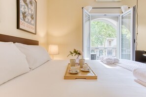 A hotel room with a bed, a tray with cups and a vase, a window with a view of buildings, and a framed picture on the wall. - Easylife - Studio with balcony in Porta Romana (Milano)