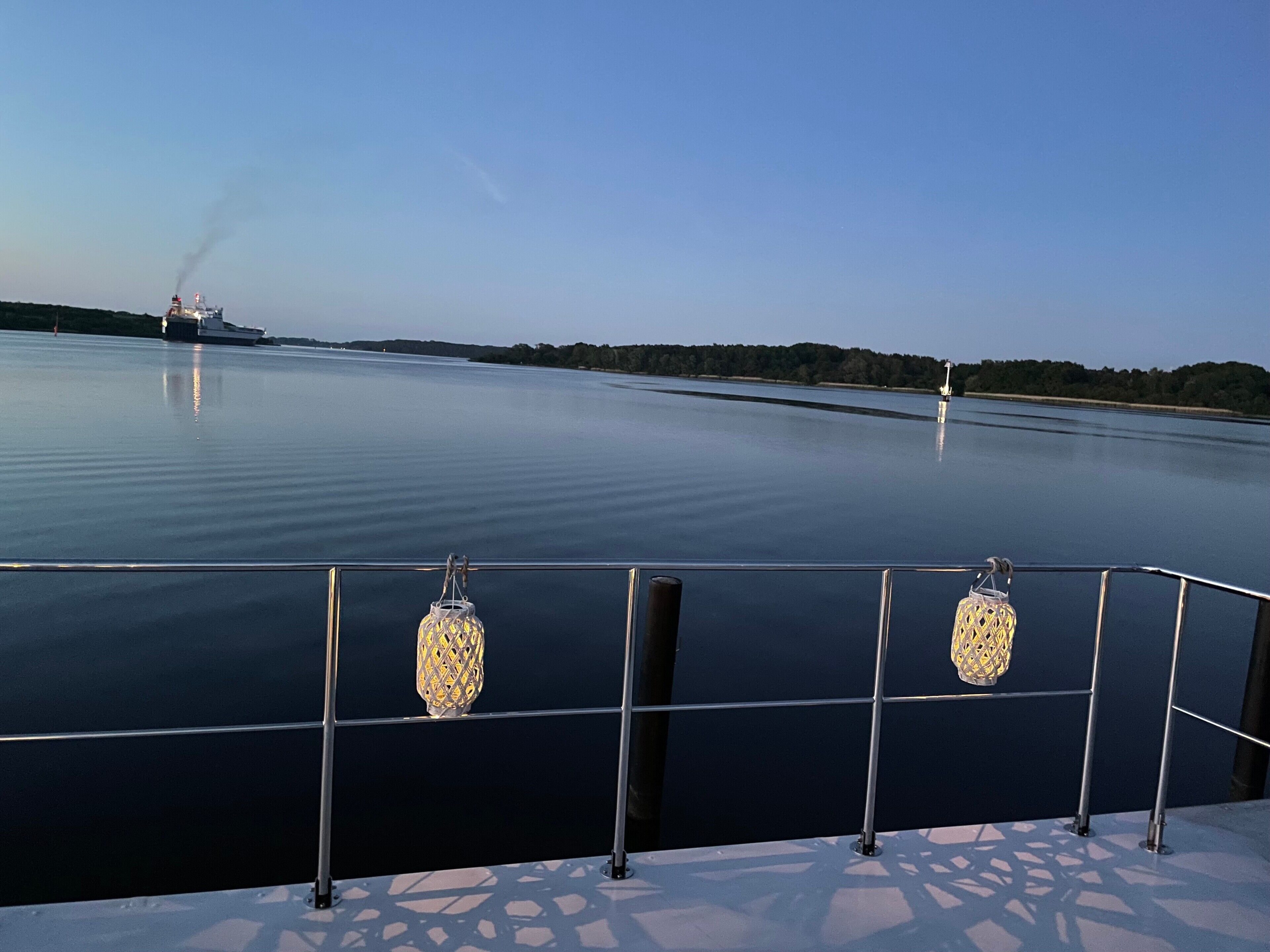 Une étendue d’eau calme, avec un bateau visible au loin, une balustrade ornée de lanternes décoratives et un ciel dégagé.