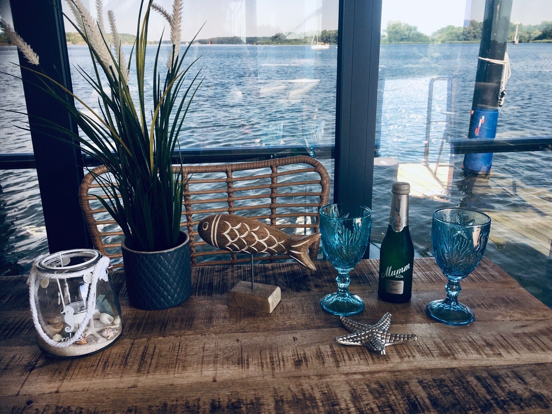 A wooden table with a potted plant, a decorative fish, a bottle of Mumm Dry, and two blue glass cups.