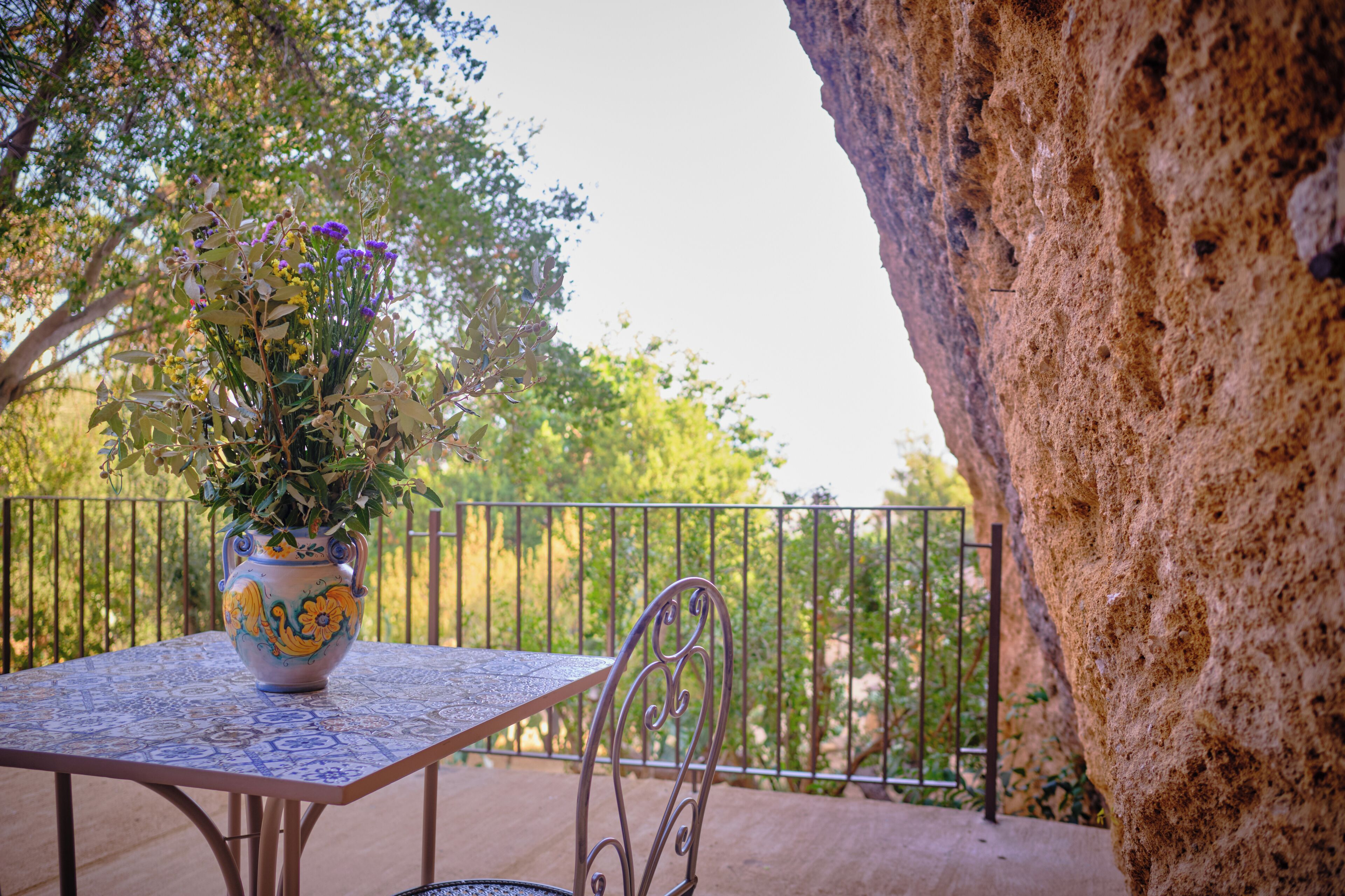 A table with a decorative vase on it, a metal chair, and a railing with greenery in the background.
