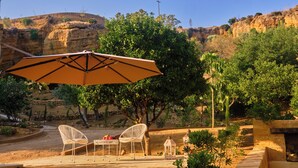 An outdoor patio with a table and chairs under a large umbrella, surrounded by greenery and a rocky landscape. - VILLA CAVETTA  (Agrigento)