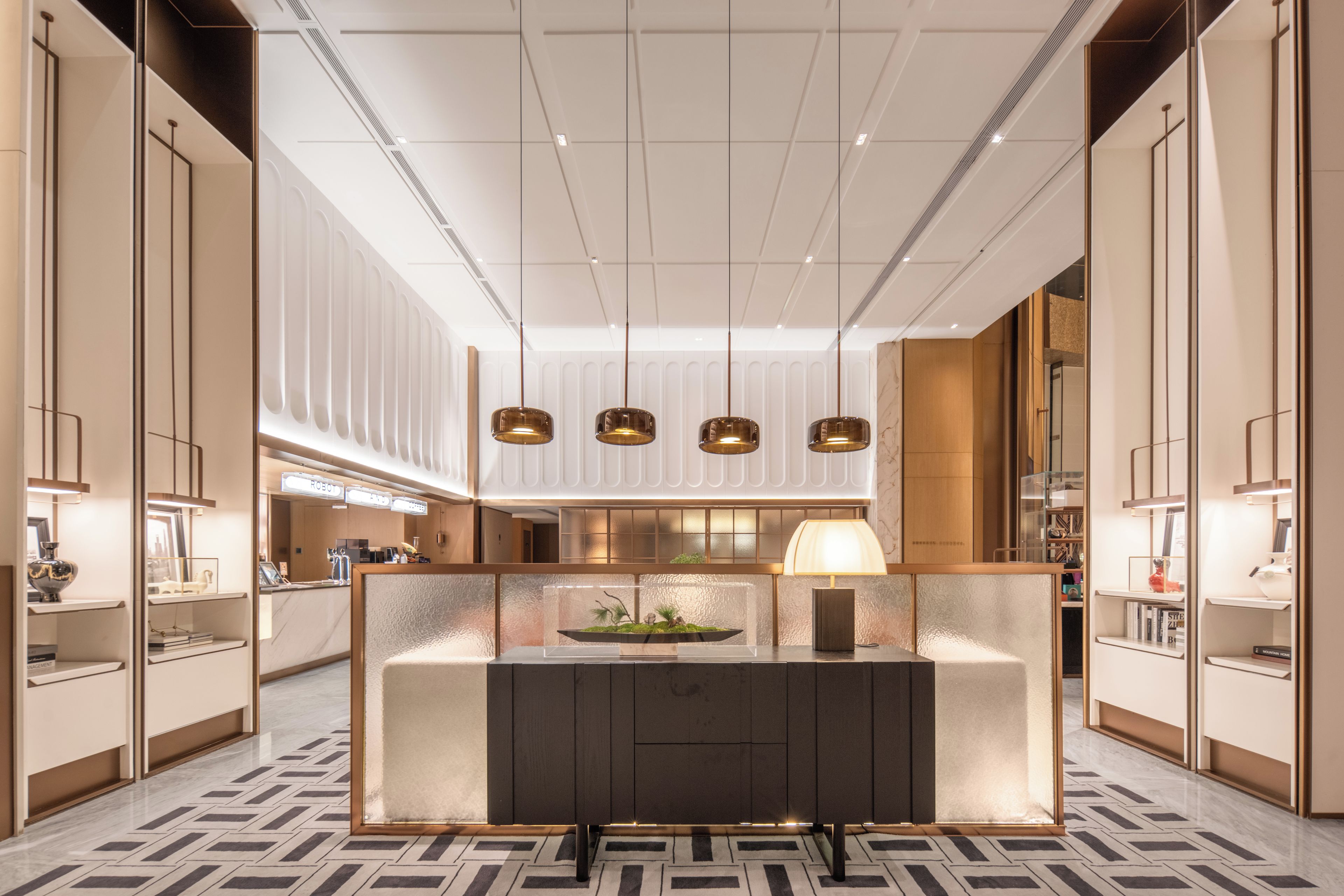 A modern hotel lobby with a central reception desk, pendant lighting, and a patterned floor.