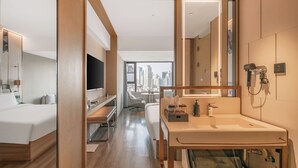 Ein modernes Hotelzimmer mit einem großen Bett, einem Fernseher, einem Schreibtisch, einem Badezimmer mit Waschbecken und Spiegel sowie Blick auf die Stadt.