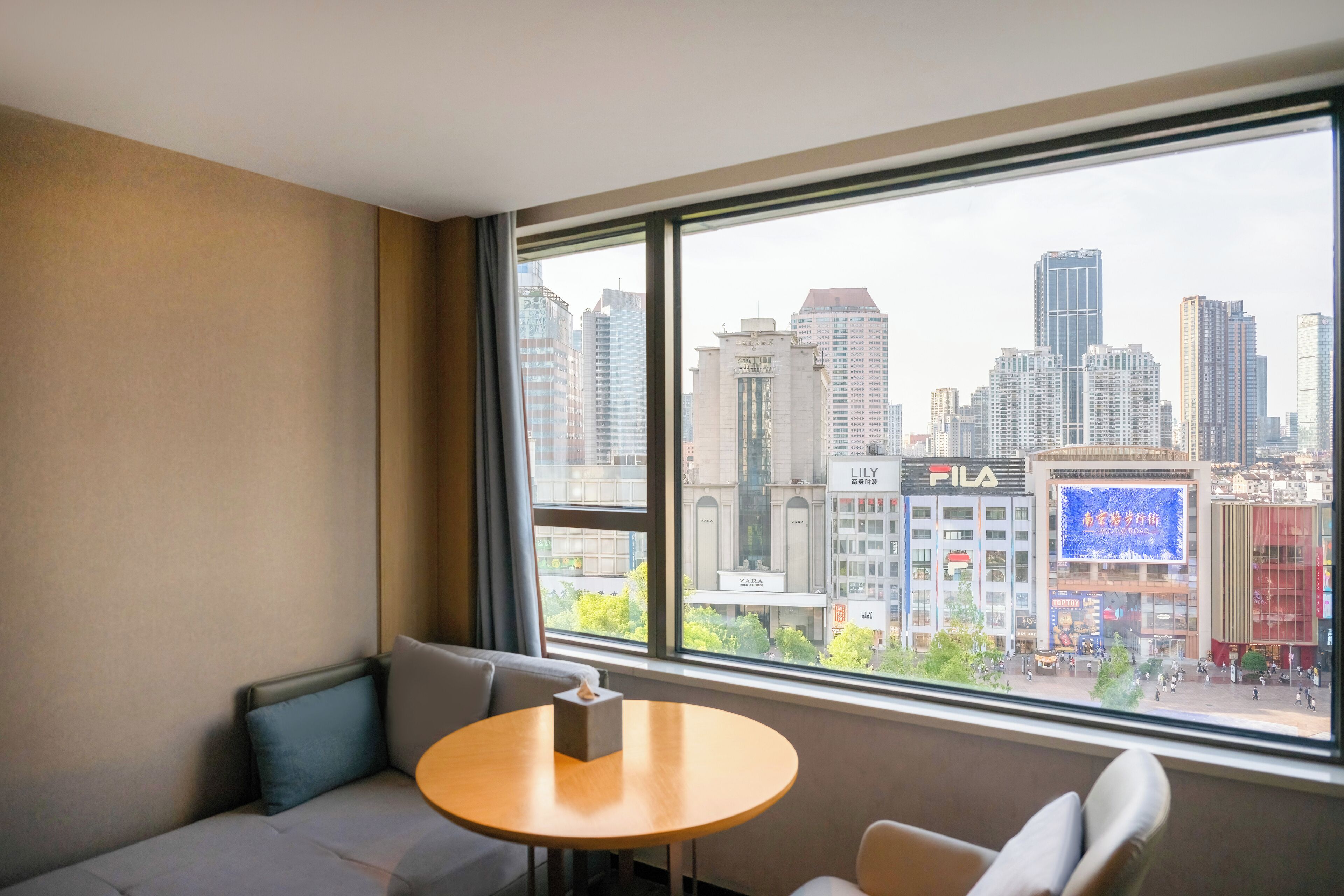 Habitación de hotel con un ventanal amplio que ofrece vistas a un paisaje urbano, incluyendo una tienda Zara.